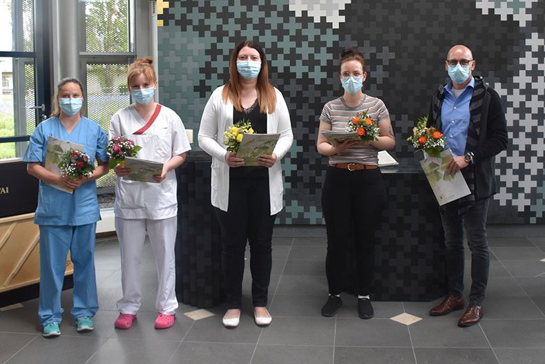 Fünf Personen mit Blumensträußen und Masken, stehen vor einer pixelartigen Wand.