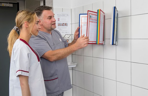 Zwei Mitarbeiter in grauer Arbeitskleidung betrachten Dokumente an der Wand.