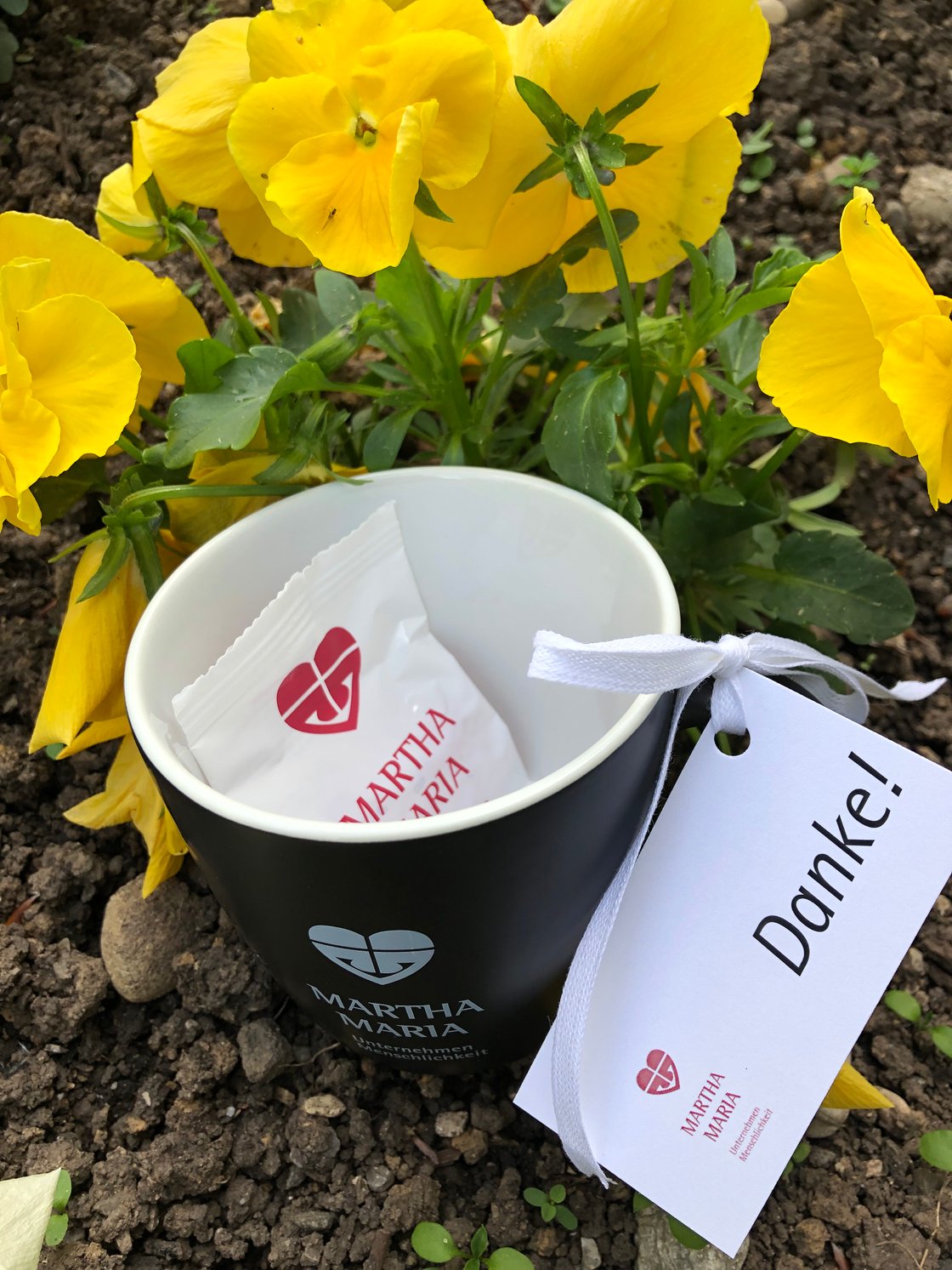 Schwarze Tasse mit 'Martha Maria'-Logo vor gelben Blumen, Dankeskarte angehängt.