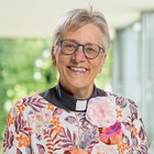 Lächelnde Frau mit Brille und Blumenkleidung vor unscharfem Grün.