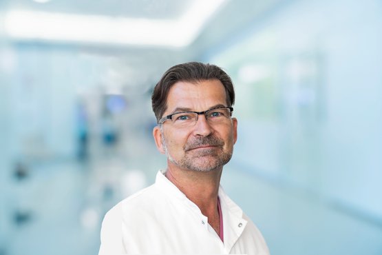 Mann mit Brille in weißem Arztkittel vor unscharfem Krankenhausflur.