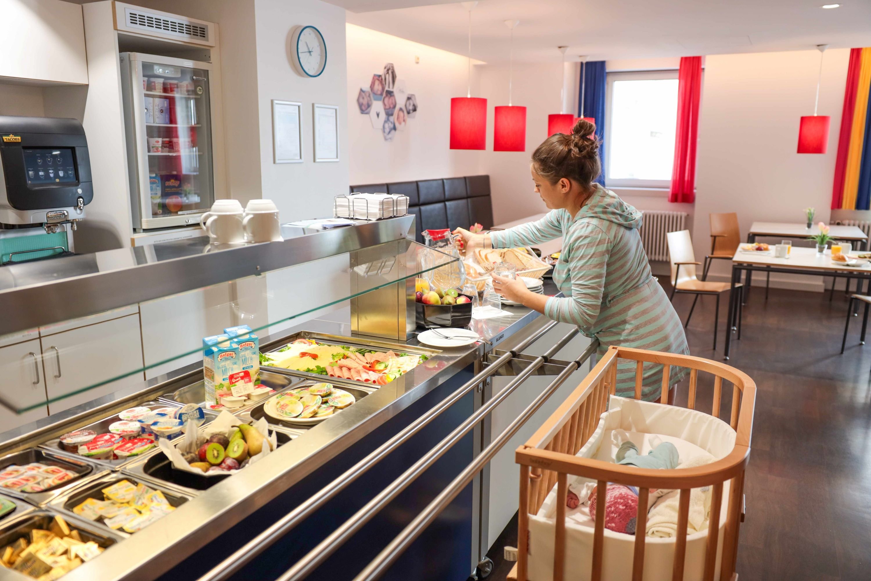 Mutter am Frühstücksbuffet im Krankenhaus mit Baby im Bettchen.