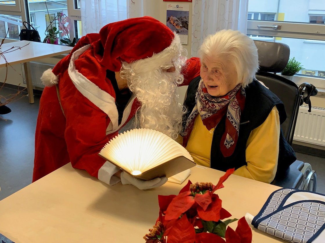 Weihnachtsmann liest Seniorin im Rollstuhl ein Buch vor.