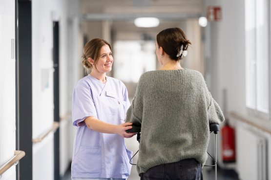 Pflegekraft unterstützt Patientin mit Gehhilfe im Krankenhausflur.