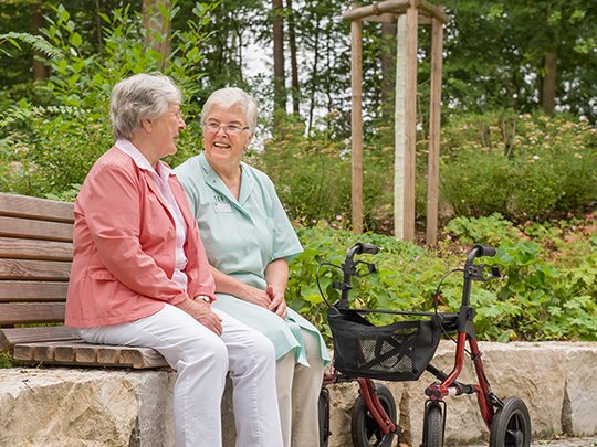 Zwei ältere Frauen auf einer Bank im Garten, eine mit Rollator.