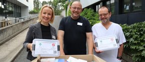 Drei Personen halten Alt-Arzneimittelsammelboxen vor einem Krankenhausgebäude.