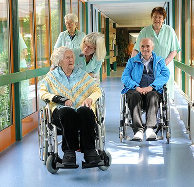 Grüne Damen und Herren begleiten Patienten im Rollstuhl im Krankenhausflur.