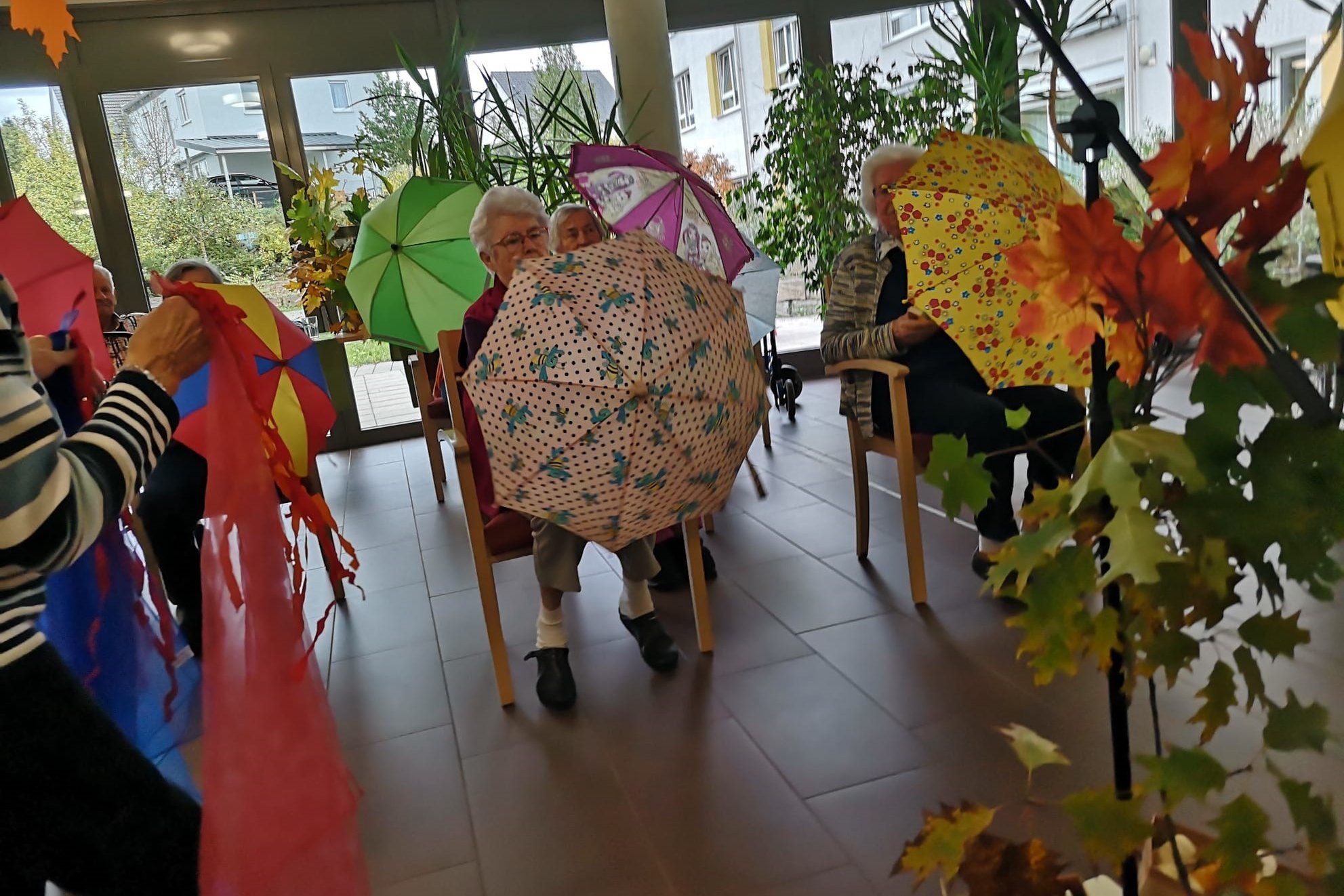 Senioren im Gemeinschaftsraum mit bunten Regenschirmen und Herbstdekoration.