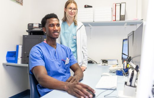 Zwei Pflegekräfte in Krankenhauskleidung arbeiten am Computer in einem Büro.
