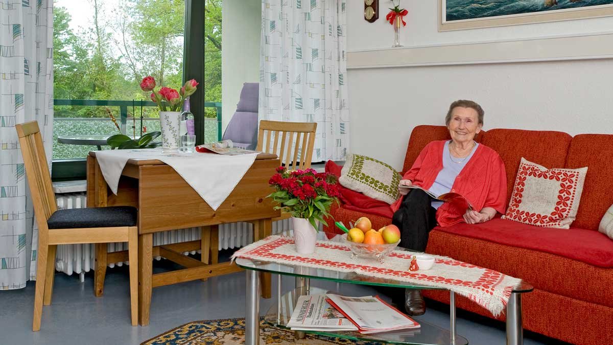 Seniorin sitzt lächelnd auf rotem Sofa in gemütlichem Wohnzimmer.