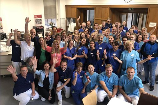 Große Gruppe lächelnder Krankenhausmitarbeiter, einige in blauer Kluft, winkend.