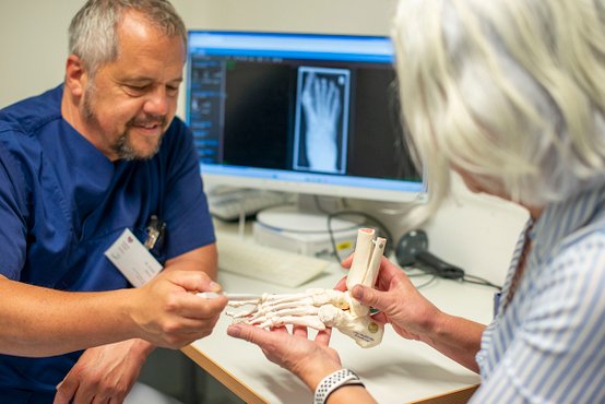 Arzt erklärt Patienten Fußmodell vor Röntgenbild am Bildschirm.