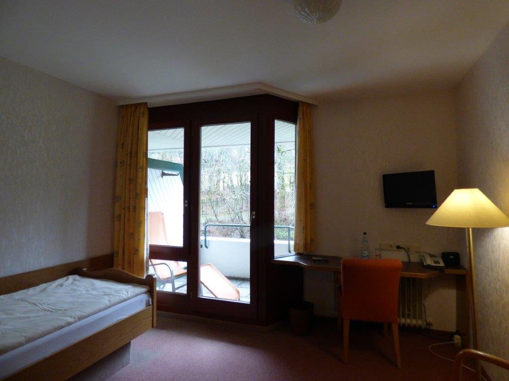 Einzelzimmer mit Bett, Schreibtisch und Balkon im Krankenhaus.