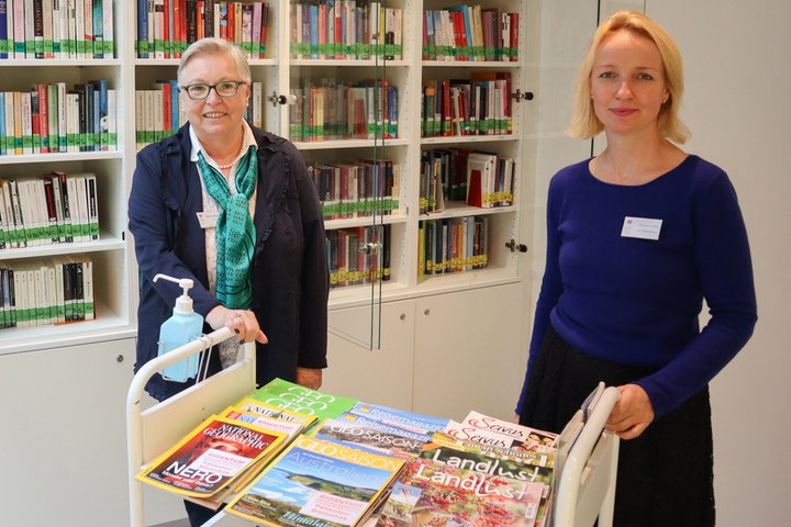 Zwei Frauen in einer Bibliothek mit Bücherregalen im Hintergrund.