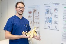 Arzt in blauer Uniform hält Hüftgelenkmodell vor medizinischen Postern.