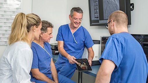 Ärzte-Team in blauer Kleidung bespricht kardiologische Befunde auf einem Tablet.