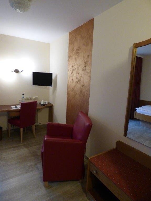 Krankenhauszimmer mit rotem Sessel, Schreibtisch und Fernseher.