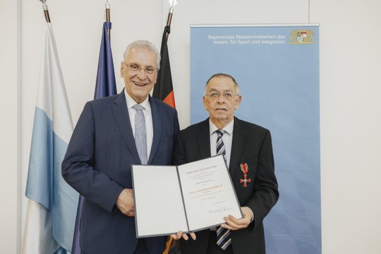Zwei Männer, einer hält Verdienstkreuz-Urkunde, vor bayerischem Ministerium.