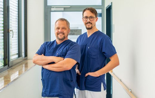 Zwei Ärzte in blauen Kitteln lächeln im Krankenhausflur.