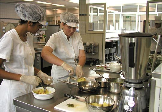 Zwei Köchinnen in weißer Kleidung bereiten Essen in Krankenhausküche zu.