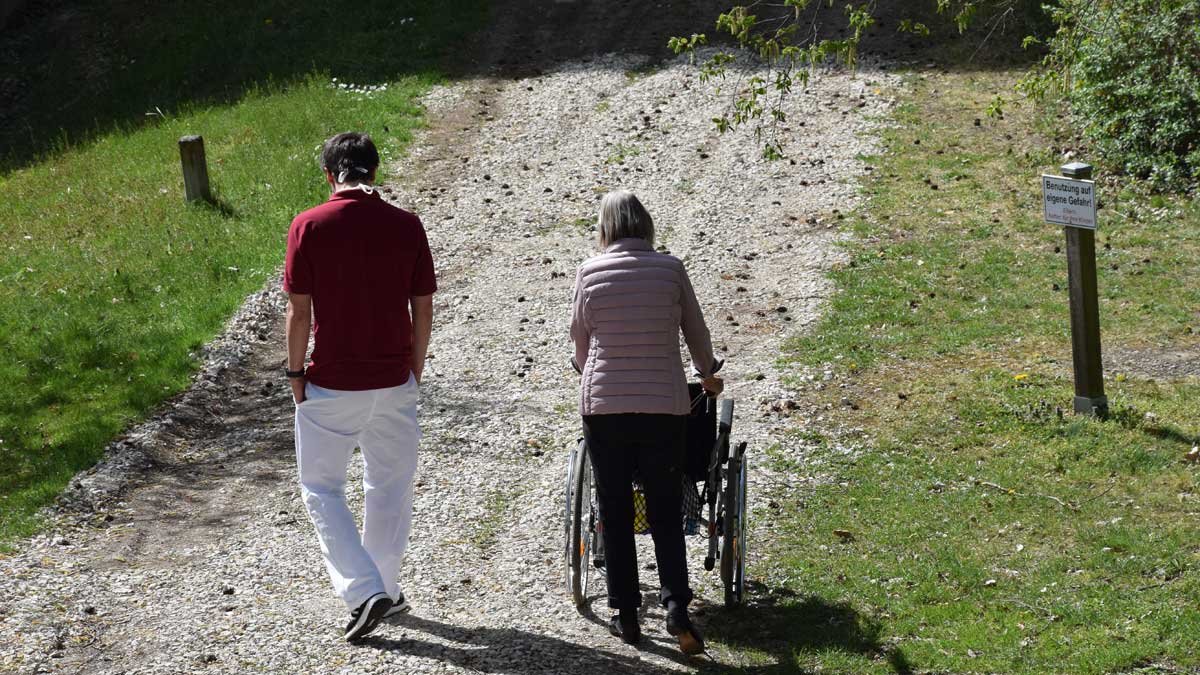 Pflegekraft begleitet Seniorin mit Rollstuhl auf Kiesweg im Grünen.