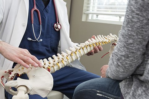 Arzt erklärt Wirbelsäulenmodell einem sitzenden Patienten in medizinischer Umgebung.