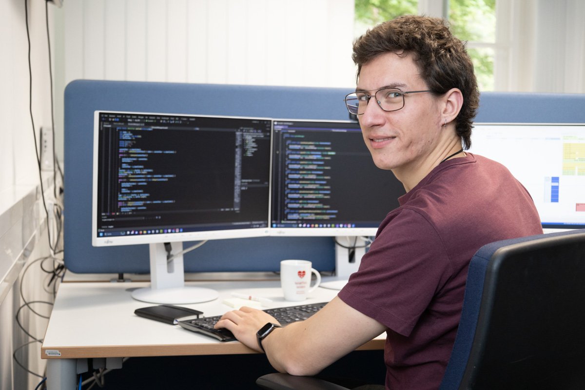 Junger Mann im Büro, arbeitet lächelnd an zwei Monitoren mit Programmiercode.