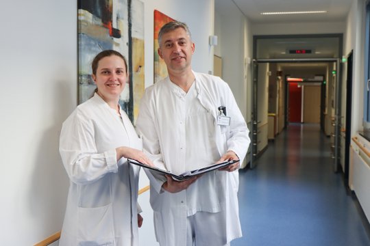 Zwei Ärzt:innen in weißen Kitteln stehen im Klinikflur und lächeln.