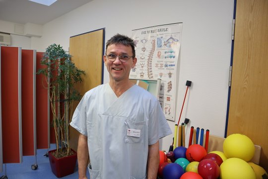 Therapeut in Behandlungsraum mit Trainingsgeräten und Poster der Wirbelsäule.