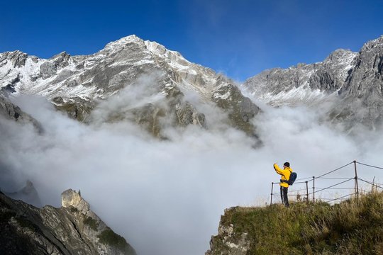 Person in gelber Jacke fotografiert Berglandschaft mit Wolken und schneebedeckten Gipfeln.