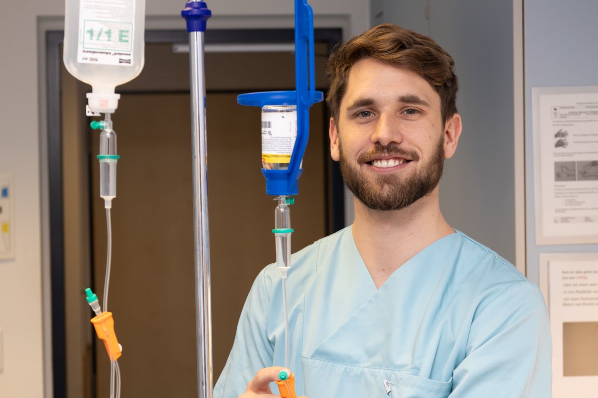 Männlicher Krankenpfleger in blauer Uniform lächelt, hält Infusionsständer im Krankenhaus.