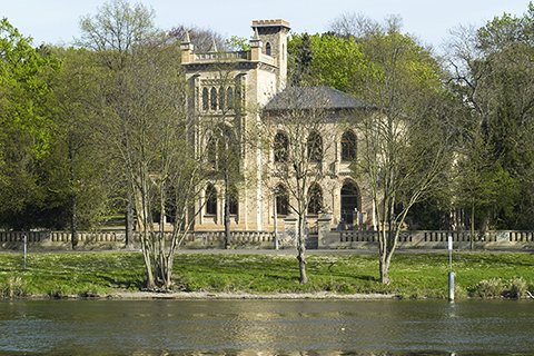 Historisches Gebäude am Flussufer, umgeben von Bäumen.