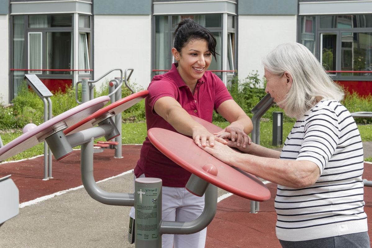 Pflegekraft hilft Seniorin bei Reha-Übung im Freien im Seniorenzentrum Stuttgart.
