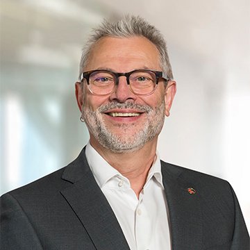 Porträt eines lächelnden Mannes in Anzug mit Brille, vor unscharfem Hintergrund.