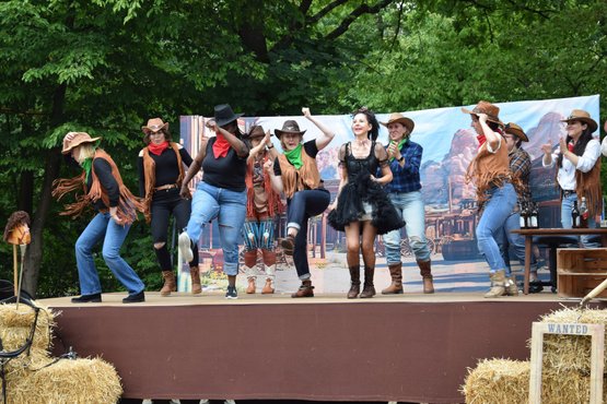 Gruppe tanzt auf Bühne in Westernkostümen vor Kulisse im Freien.