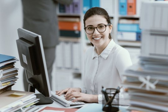Lächelnde Bürokraft am Computer, umgeben von Aktenordnern im Büro.