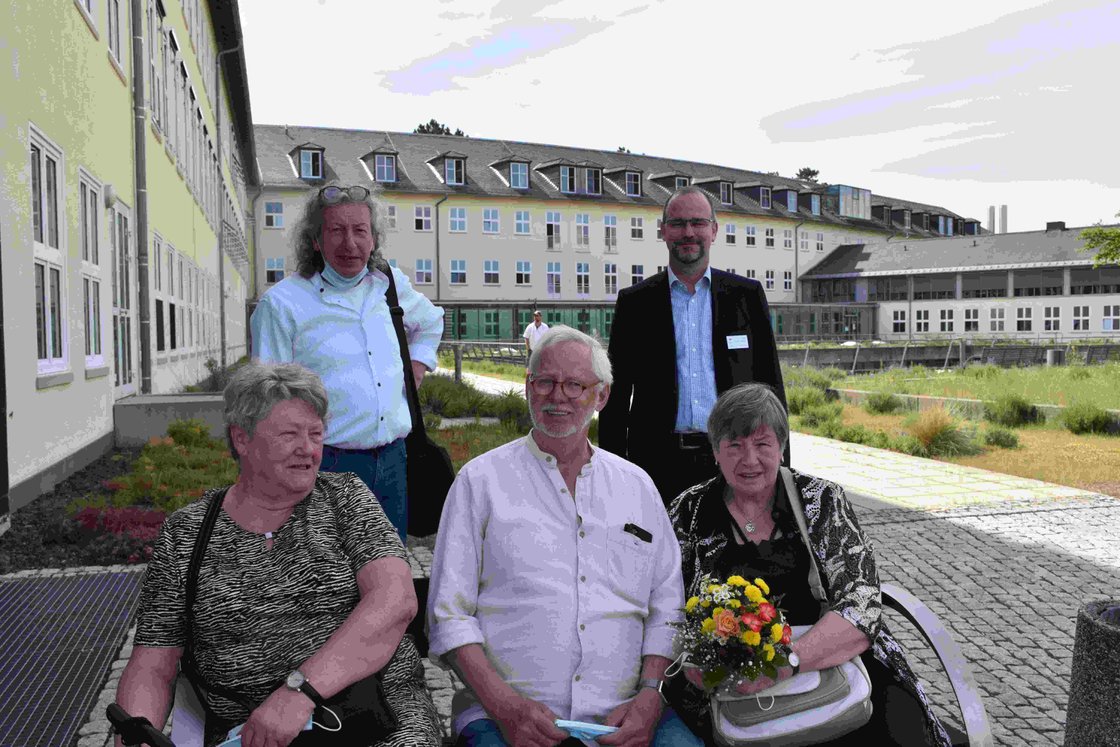 Gruppe von Senioren vor einem modernen Gebäude, zwei Frauen mit Blumenstrauß.