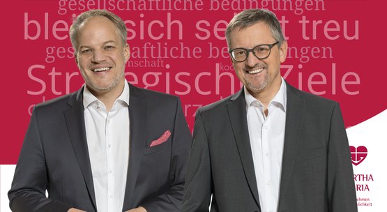 Zwei lächelnde Männer in Anzügen vor rotem Hintergrund mit Text.
