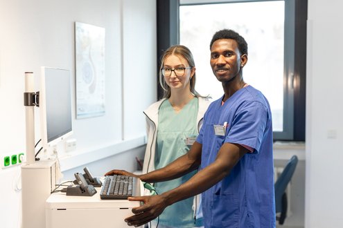 Zwei medizinische Fachkräfte in Krankenhauskleidung lächeln vor einem Computer.