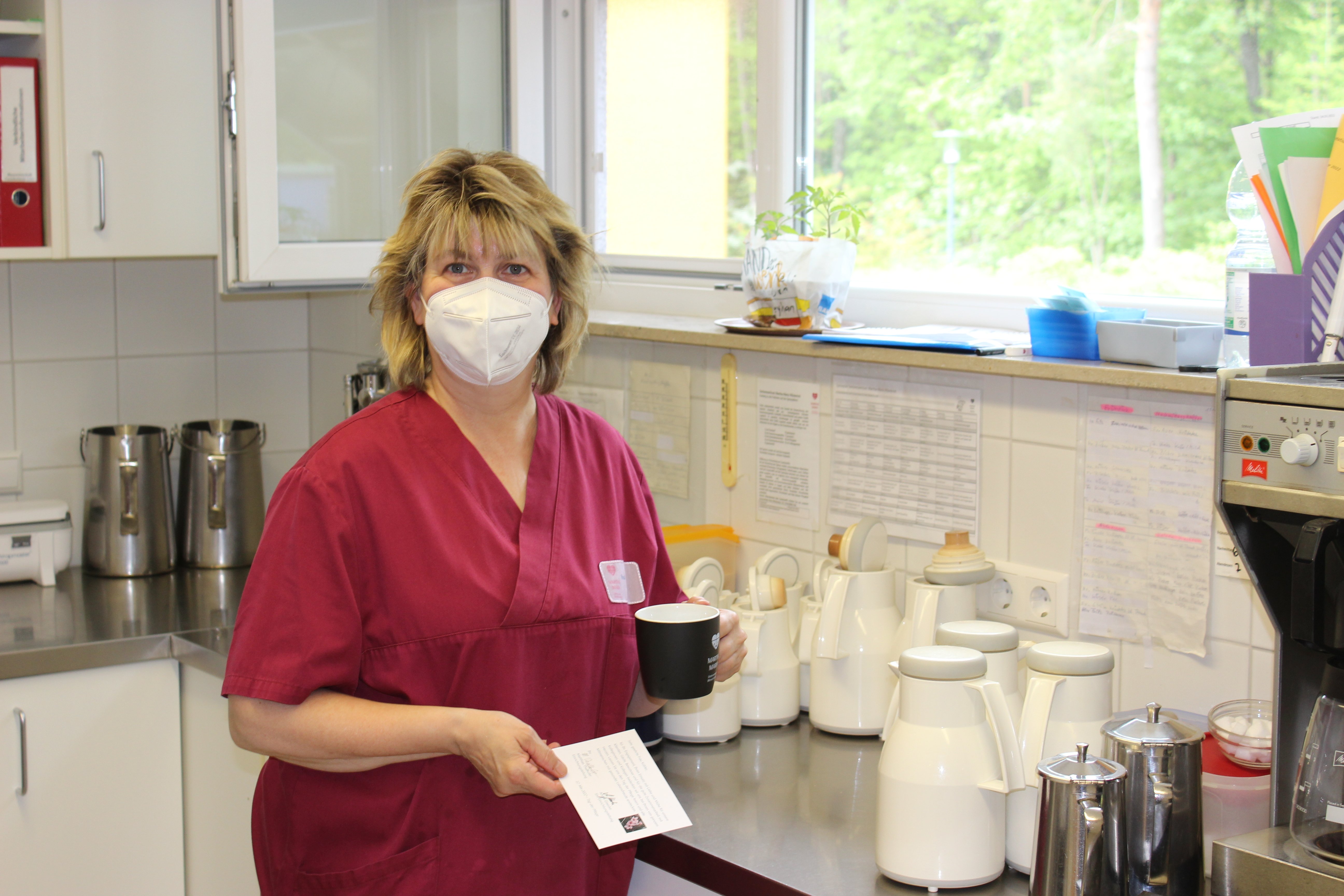 Pflegekraft in roter Uniform mit Maske in Krankenhausküche mit Kaffeekannen.