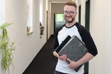 Mann mit Ordner lächelt im Büroflur, Pflanzen im Hintergrund.