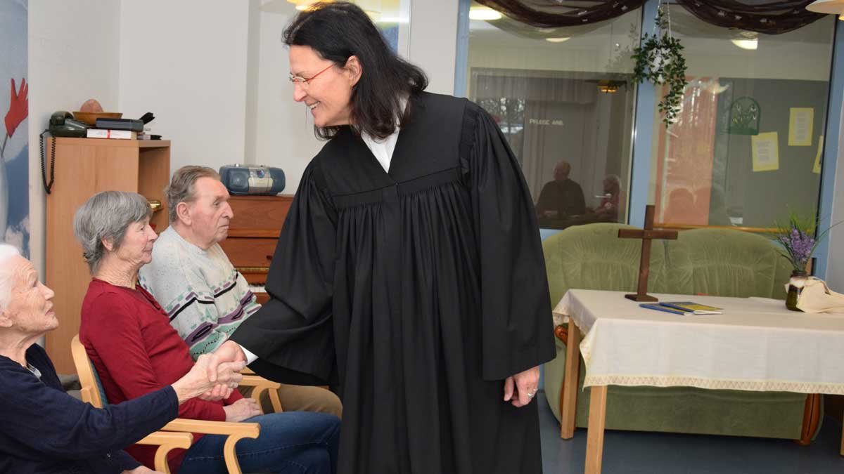 Pfarrer begrüßt Seniorin im Seniorenzentrum Nürnberg, daneben zwei sitzende Senioren.