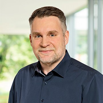 Porträt eines lächelnden Mannes in dunklem Hemd vor verschwommenem Hintergrund.