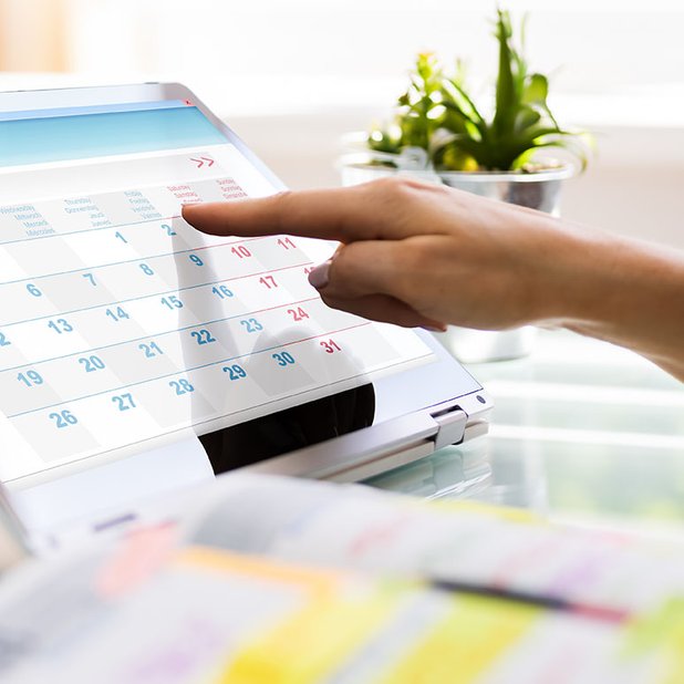 Finger tippt auf Tablet-Kalender, grünes Pflanze im Hintergrund.