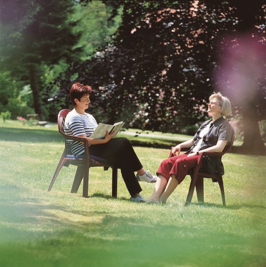 Zwei Frauen sitzen im Garten, eine liest vor, entspannte Atmosphäre.