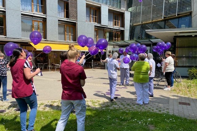 Pflegepersonal lässt lila Luftballons vor einem modernen Gebäude steigen.