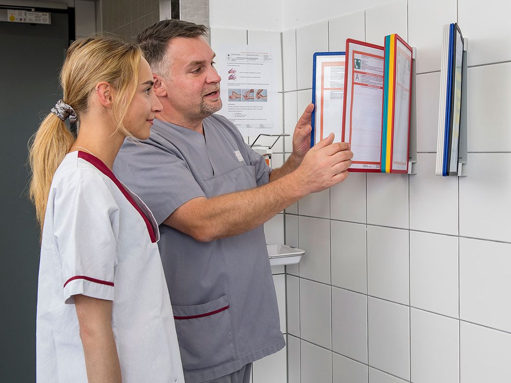 Zwei Mitarbeiter in grauer Arbeitskleidung betrachten Dokumente an der Wand.