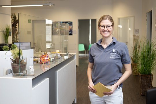 Lächelnde Mitarbeiterin in Martha-Maria-Poloshirt an Empfangstresen stehend.