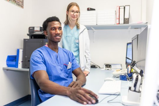 Zwei Pflegekräfte in Krankenhauskleidung arbeiten am Computer in einem Büro.