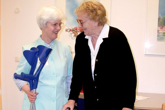 Grüne Dame hilft älterer Frau mit Gehhilfe im Krankenhaus.